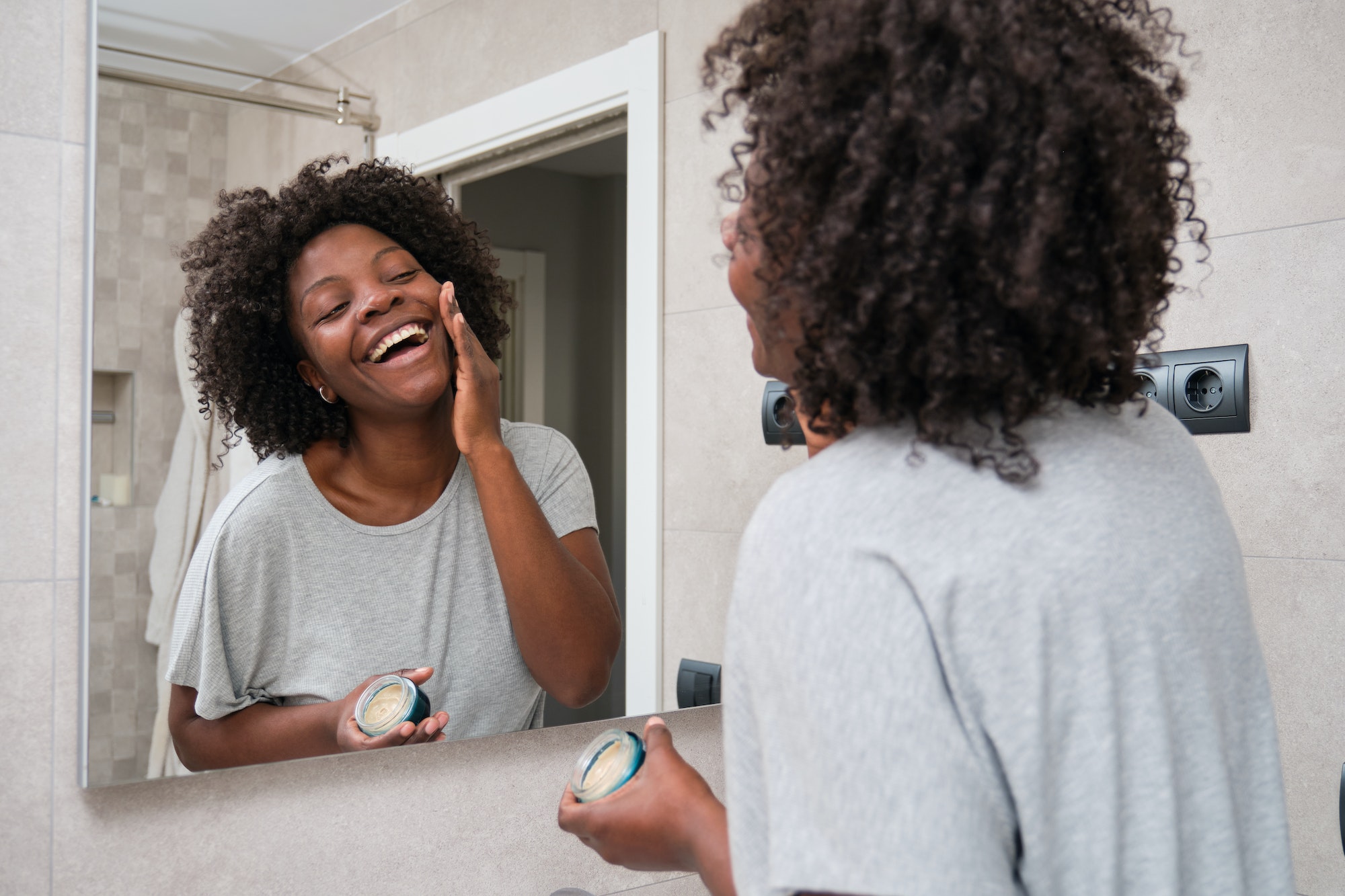 Smiling african woman applies eco-friendly facial cream in bathroom.